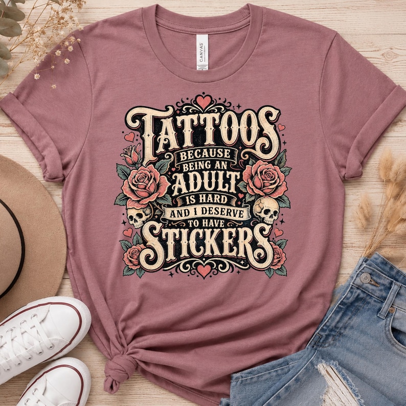 May include: Dusty rose t-shirt with a graphic that reads "Tattoos because being an adult is hard and I deserve to have stickers." The design includes roses, skulls, and hearts. The shirt is made of a soft material.