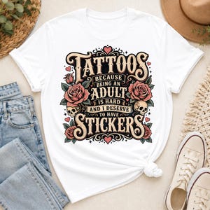 May include: White t-shirt featuring a graphic design with the words "Tattoos" and "Stickers." The design includes roses, skulls, and the phrase "Because being an adult is hard and I deserve to have." The shirt is knotted at the waist.