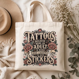 May include: Cream-colored tote bag with the text "Tattoos because being an adult is hard and I deserve to have stickers." The design includes roses, skulls, and hearts in a vintage tattoo style.