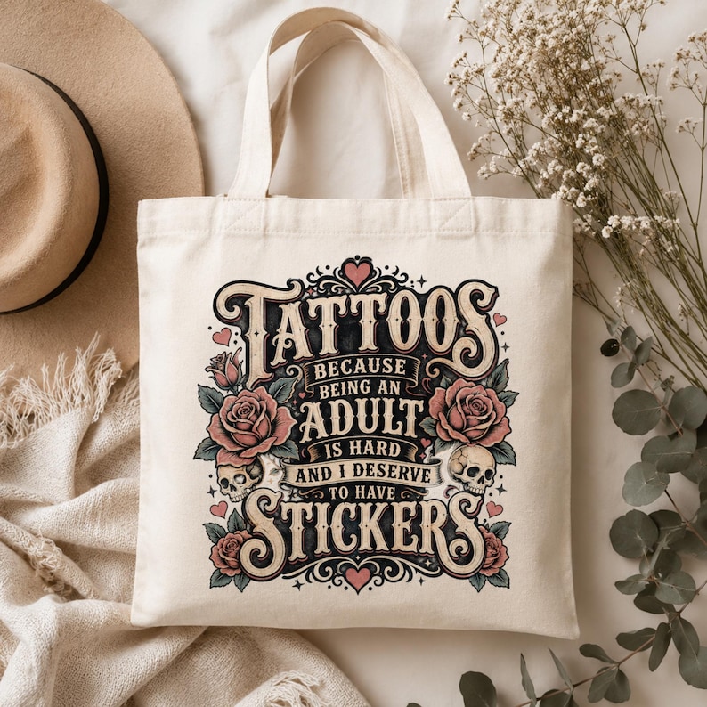 May include: Cream-colored tote bag with the text "Tattoos because being an adult is hard and I deserve to have stickers." The design includes roses, skulls, and hearts in a vintage tattoo style.