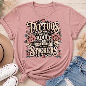 May include: Dusty rose t-shirt with a graphic that reads "Tattoos because being an adult is hard and I deserve to have stickers." The design includes roses, skulls, and decorative elements.
