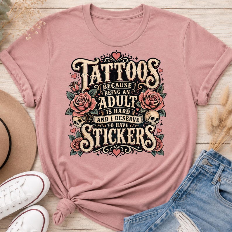 May include: Dusty rose t-shirt with a graphic that reads "Tattoos because being an adult is hard and I deserve to have stickers." The design includes roses, skulls, and decorative elements.