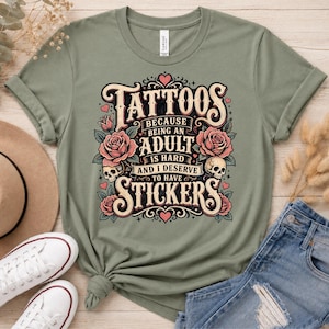 May include: Sage green t-shirt with the text "Tattoos because being an adult is hard and I deserve to have stickers." The graphic includes roses, skulls, and hearts in a vintage style. The shirt is paired with denim jeans and white sneakers.