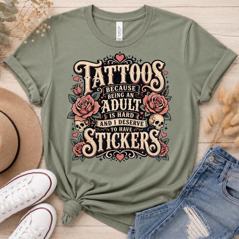 May include: Sage green t-shirt with the text "Tattoos because being an adult is hard and I deserve to have stickers." The graphic includes roses, skulls, and hearts in a vintage style. The shirt is paired with denim jeans and white sneakers.