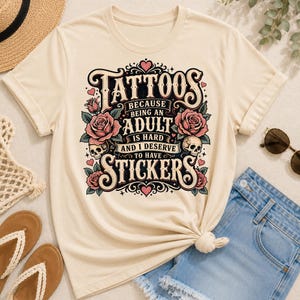 May include: Cream-colored t-shirt featuring the text "Tattoos because being an adult is hard and I deserve to have stickers." The design includes roses, skulls, and decorative flourishes in black, pink, and green.