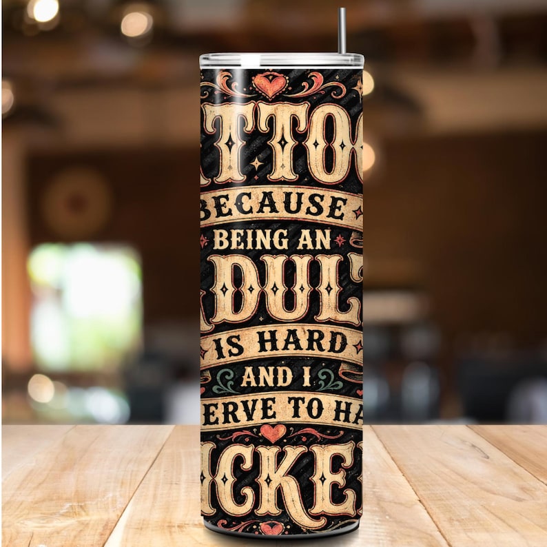 K&ouml;nnte beinhalten: Ein schwarz-becher mit Strohhalm, mit dem Text "Tattoos because being an adult is hard and I deserve to have cake." Der Text ist in einer Vintage-Schrift mit Herzakzenten gehalten. Der Becher steht auf einer Holzoberfl&auml;che.