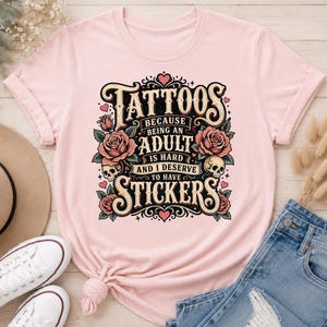 May include: Light pink t-shirt with a graphic design. The design features the word "Tattoos" in a vintage font, along with roses, skulls, and the text "Because being an adult is hard and I deserve to have stickers."