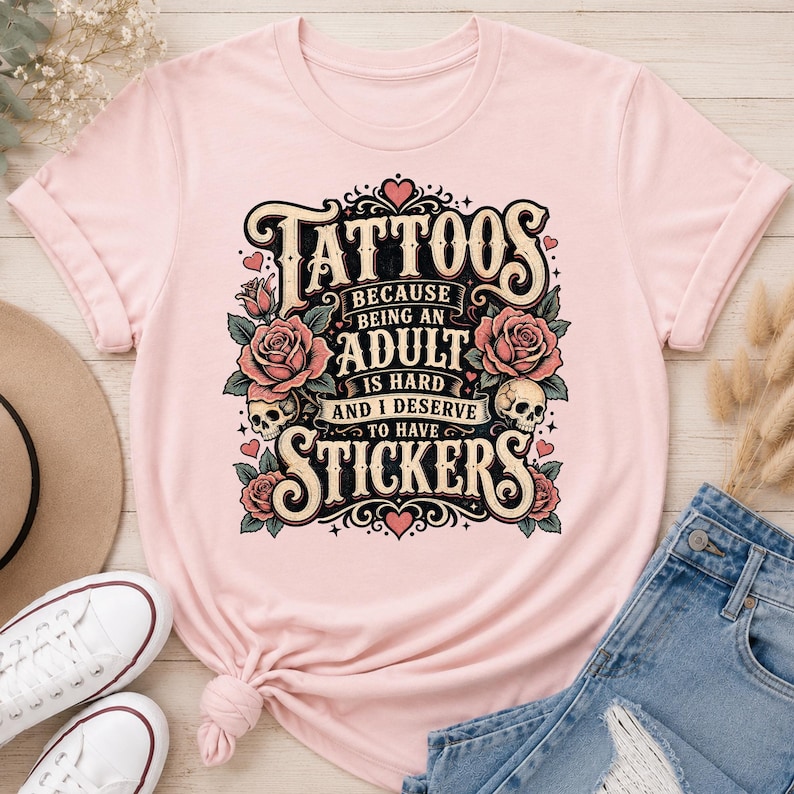 May include: Light pink t-shirt with a graphic design. The design features the word "Tattoos" in a vintage font, along with roses, skulls, and the text "Because being an adult is hard and I deserve to have stickers."