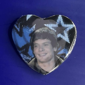 May include: Heart-shaped button with a photograph of a smiling person. The background features a blue and black star and spray paint design. The button is likely a collectible or a piece of memorabilia.