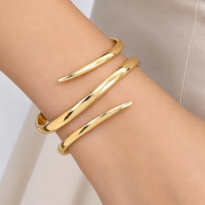 Può includere: Un bracciale a fascia in metallo color oro lucido con un design a spirale che avvolge il polso. Il bracciale presenta una superficie liscia e riflettente con estremità affusolate che si curvano verso l'esterno in una forma a spirale.