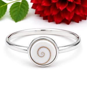 Puede incluir: Un anillo de plata con un engaste circular que contiene una incrustación de concha de ojo de shiva blanca. La concha muestra un patrón de espiral marrón natural centrado en la banda de metal pulido. El anillo tiene un acabado liso y brillante.