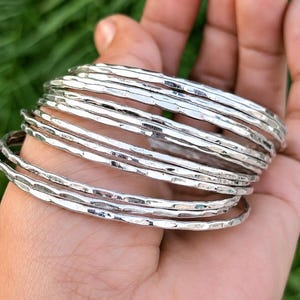 May include: A set of multiple thin, circular silver-colored bangles. Each bangle features a distinct hammered, textured surface that reflects light, creating a series of uneven, metallic ridges along the circumference of the individual bracelets.