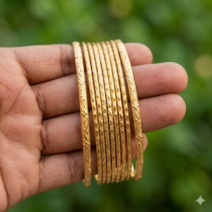 May include: A collection of gold bangles, each adorned with detailed floral and textured patterns. The bangles are held in a hand, set against a soft green backdrop. The jewelry's warm gold tone and delicate designs are highlighted. The bangles are likely for adornment.