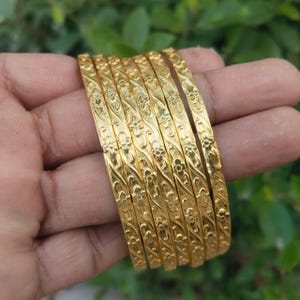 Op de afbeelding: Een verzameling gouden armbanden met bloempatronen. De armbanden zijn op elkaar gestapeld en tonen hun ingewikkelde ontwerpen. De goudkleur is helder en glanzend en reflecteert licht. De achtergrond is een vage groene kleur.