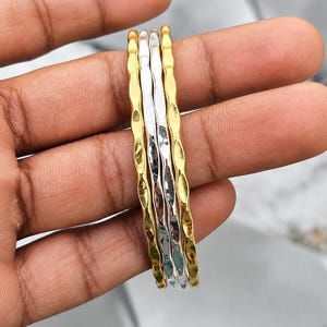 Może przedstawiać: Zestaw czterech cienkich metalowych bransoletek typu bangle o falistej, młotkowanej fakturze. Stos składa się z dwóch złotych bransoletek na zewnątrz i dwóch srebrnych w środku, wszystkie z polerowanym, odblaskowym wykończeniem.