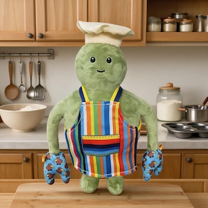 Pode incluir: Uma criatura de pelúcia verde usando um chapéu de chef branco e um avental de listras verticais coloridas com um bolso frontal. A criatura tem dois braços com luvas de forno azuis com um padrão de pássaros kiwi marrons, cabeça redonda e rosto.