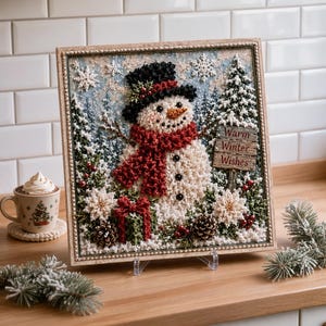 May include: A square framed wall art piece featuring a textured 3D snowman wearing a black top hat and a red scarf. The scene includes pine trees, snowflakes, pinecones, and a wooden sign with the text Warm Winter Wishes in red lettering.