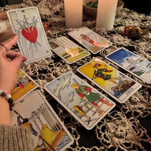 May include: A collection of tarot cards, featuring various illustrations and symbols, are arranged on a table. The cards depict scenes with swords, figures, and a heart pierced by swords. Two lit candles and a decorative cloth complete the scene.