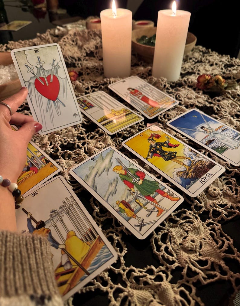 May include: A collection of tarot cards, featuring various illustrations and symbols, are arranged on a table. The cards depict scenes with swords, figures, and a heart pierced by swords. Two lit candles and a decorative cloth complete the scene.