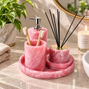 Puede incluir: Un juego de accesorios de baño de mármol rosa y blanco sobre una bandeja circular a juego. El conjunto incluye un dispensador de jabón con bomba plateada, un portacepillos con dos cepillos de bambú y un difusor con varillas negras.
