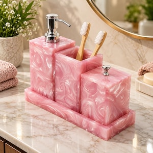 Può includere: Un set da bagno composto da un dispenser di sapone in resina marmorizzata rosa con erogatore argentato, un portaspazzolini abbinato contenente due spazzolini in bambù e un contenitore con coperchio, il tutto su un vassoio rettangolare in resina.