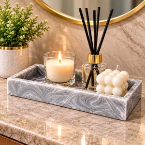 Peut inclure: Un plateau rectangulaire en marbre gris et blanc contenant un bougeoir en verre transparent avec une bougie blanche allumée, un diffuseur à roseaux carré avec des bâtonnets noirs et un col doré, et une bougie pilier à bulles.