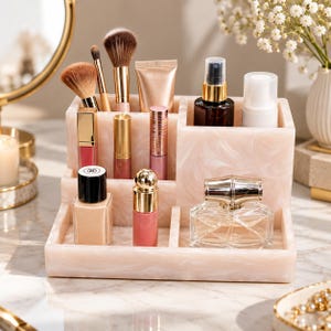 Könnte beinhalten: Ein mehrstufiger Make-up-Organizer aus Acryl mit hellrosa Marmormuster, der verschiedene Kosmetika enthält, darunter Make-up-Pinsel, Lippenstifttuben, Foundation und einen Glasparfümflakon mit goldenem Verschluss in separaten Fächern.