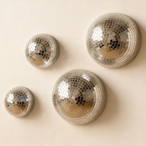 Op de afbeelding: Vier zilveren discobol-decoraties van verschillende formaten zijn op een lichtgekleurde muur gemonteerd. Elke bal is bedekt met kleine, reflecterende spiegeltegels, waardoor een glinsterend effect ontstaat. De opstelling suggereert een decoratief thema, geschikt voor feesten of interieurontwerp.