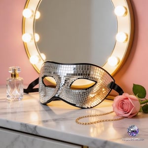 May include: A silver disco ball mask with black trim and black ties rests on a marble surface. A gold-framed mirror with lights, a perfume bottle, a pink rose, and a gold chain are also present.