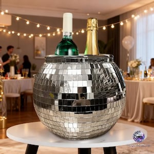 Könnte beinhalten: Ein silberner Weinkühler in Form einer Discokugel mit zwei Flaschen, eine grüne und eine goldene. Der Kühler steht auf einem weißen Tisch mit schwarzen Beinen. Der Hintergrund zeigt eine Party mit Lichterketten und Menschen.