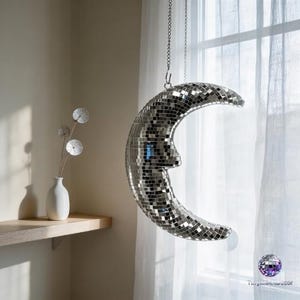 May include: A crescent moon-shaped disco ball hangs from a chain in front of a window. The silver-tiled surface reflects light. A white vase with decorative stems sits on a wooden shelf. The room has neutral-colored walls and a sheer white curtain.