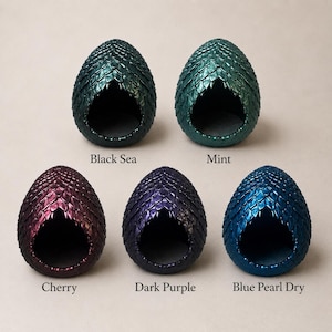 Op de afbeelding: Vijf eivormige houders met een gestructureerd schubpatroon. De kleuren zijn gelabeld als Black Sea, Mint, Cherry, Dark Purple en Blue Pearl Dry. Elk object heeft een grote, uitgeholde opening aan de voorzijde die een gladde binnenkant onthult.