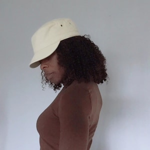 May include: A cream-coloured baseball cap with a structured crown and a curved brim, featuring small ventilation eyelets on the side panels. The cap is worn with a long-sleeved brown shirt that has a fitted silhouette and a high neckline.