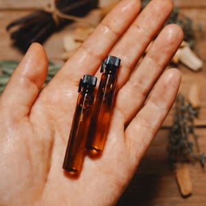 Puede incluir: Dos pequeños viales de vidrio ámbar con tapas negras se sostienen en una mano abierta. Los viales contienen un líquido transparente. El fondo incluye una superficie de madera y hierbas secas, lo que sugiere un tema natural o de aromaterapia.