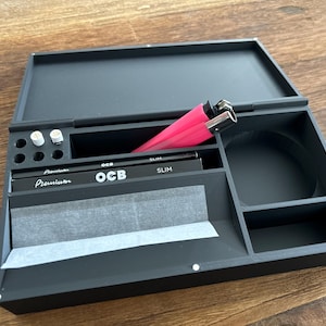 Könnte beinhalten: Eine schwarze Holz-Rollschale mit mehreren Fächern. Sie enthält eine Packung OCB Slim Zigarettenpapier, zwei Feuerzeuge und zwei weiße zylindrische Gegenstände. Die Schale ist geöffnet und zeigt ihren Inhalt.