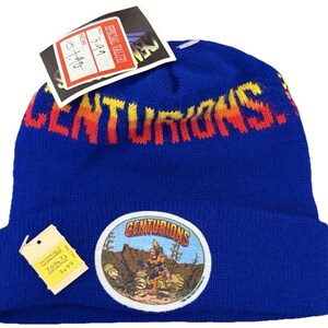 May include: A blue knit beanie with the word "CENTURIONS" in orange and yellow across the top. A circular patch with the word "CENTURIONS" and a cartoon graphic is on the front. The beanie has a folded brim and a price tag attached.