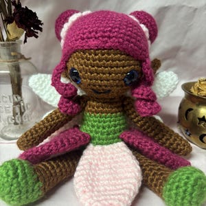 May include: A handmade crochet fairy doll with a brown face, dark pink hair, and blue eyes. The doll has white wings, brown arms and legs, and a pink, green, and white outfit. The doll is a whimsical, handcrafted item.