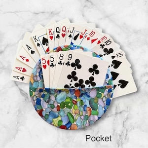 May include: A circular fabric card holder featuring a colorful pattern of smooth, rounded stones in shades of blue, green, and orange. The holder contains a fanned-out deck of standard playing cards, with the nine of clubs visible in the front.