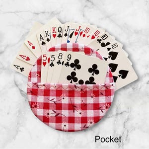 May include: A circular fabric card holder featuring a red and white gingham pattern with small floral accents. The holder contains a fanned arrangement of standard playing cards, including various suits and numbers, with the word Pocket printed below.