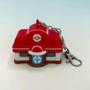 May include: A red and white silicone keychain shaped like a building with a rounded roof, featuring a central blue door with a circular emblem, a white Poké Ball icon above the entrance, and a silver metal chain with a lobster claw clasp.