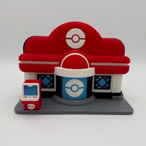 Pode incluir: Um modelo de edifício vermelho e branco com um logótipo circular azul e branco. O modelo tem um telhado vermelho e um pequeno quiosque vermelho. O edifício tem uma base cinzenta e é provavelmente um brinquedo ou um objeto de coleção.
