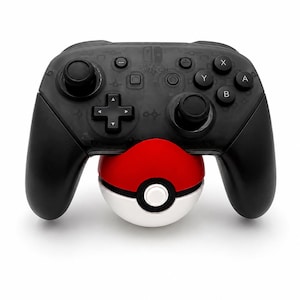 Puede incluir: Un mando Nintendo Switch Pro negro con un accesorio Pokeball rojo y blanco. El mando tiene botones y joysticks negros, con una cruceta y otros botones de funci&oacute;n. La Pokeball es roja y blanca con un bot&oacute;n blanco.