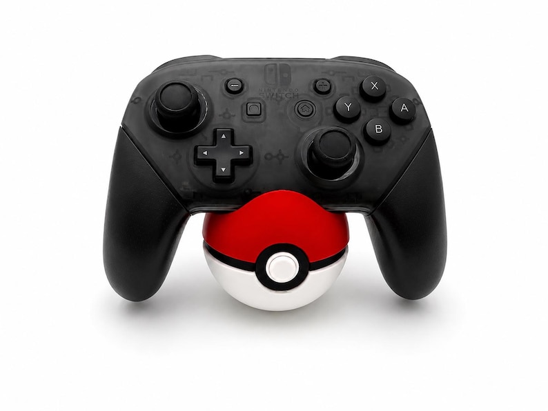 Puede incluir: Un mando Nintendo Switch Pro negro con un accesorio Pokeball rojo y blanco. El mando tiene botones y joysticks negros, con una cruceta y otros botones de funci&oacute;n. La Pokeball es roja y blanca con un bot&oacute;n blanco.