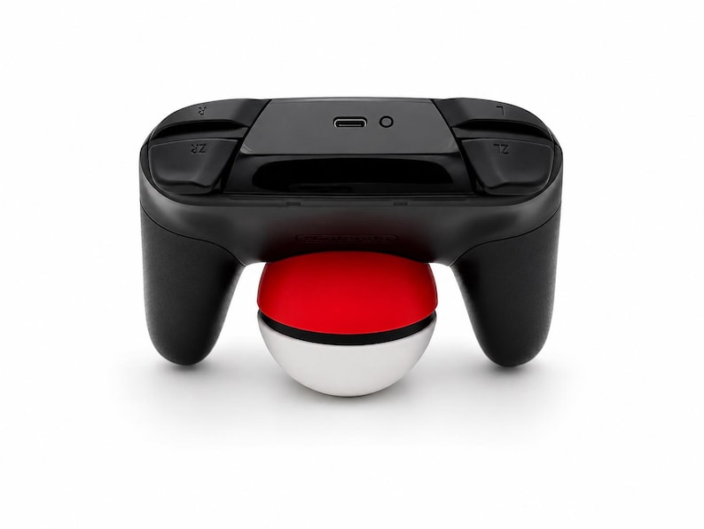 Puede incluir: Un mando de videojuegos negro con una base de Pok&eacute; Ball roja, blanca y negra. El mando tiene botones etiquetados ZL y ZR. La Pok&eacute; Ball tiene aproximadamente 7,6 cm de di&aacute;metro.