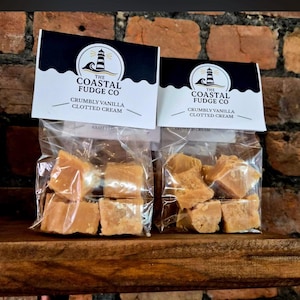 Op de afbeelding: Twee transparante zakjes gevuld met kruimelige vanille clotted cream fudge. Elk zakje heeft een label met de tekst "THE COASTAL FUDGE CO" en "CRUMBLY VANILLA CLOTTED CREAM". De fudge is lichtbruin.