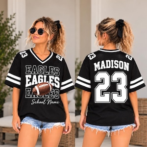 May include: A black V-neck jersey with white stripes on the sleeves and the number 23 on the shoulders. The front features the text EAGLES and a football graphic with the text School Name. The back displays the name MADISON and the number 23.