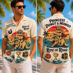 Könnte beinhalten: Beige Hemd mit Knöpfen und einer farbenfrohen Grafik einer Katze auf einem Dinosaurier, mit dem Text "Princess Donut & Mongo" und "Ride or Roar". Das Hemd hat ein tropisches Thema mit Palmen und anderen Illustrationen.