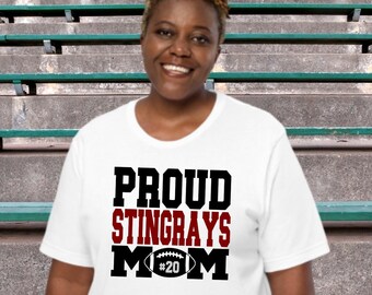 May include: White t-shirt with black text that reads "Proud Stingrays Mom" and a red number 20 inside a black football graphic.