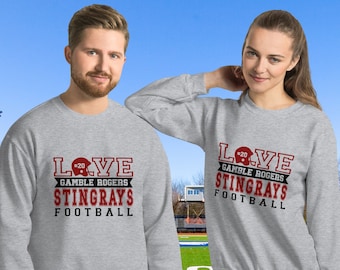 May include: Two gray sweatshirts with the text "LOVE GAMBLE ROGERS STINGRAYS FOOTBALL" printed on the front. The text is in red and white. The sweatshirts are being modeled by two people standing on a football field.