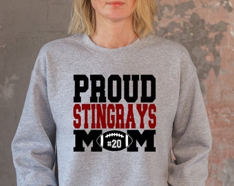 May include: A light gray sweatshirt with the words "PROUD STINGRAYS MOM #20" in black and red lettering. The design includes a football graphic. The sweatshirt is a casual, comfortable style.
